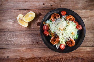 Appetizing Caesar salad with shrimps in a black plate on a wooden table next to a lemon. The concept of healthy food and serving