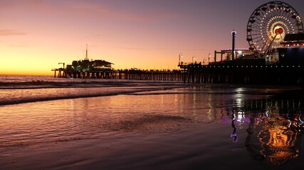 Twilight waves against classic illuminated ferris wheel, amusement park on pier in Santa Monica pacific ocean beach resort. Summertime iconic symbol of California glowing in dusk, Los Angeles, CA USA.