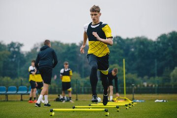 Obraz premium Teenager Boy Soccer Player in Training. Young Soccer Players at Practice Session with Coach. Footballer Jumping Over Hurdles. Boys Running Youth Agility Ladder Drills. Soccer Ladder Exercises