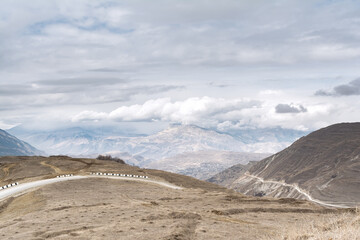 View of the alpine road against the background of the mountain peaks of a gloomy sky with clouds and dry grass of sand color. The concept of landscape, travel.