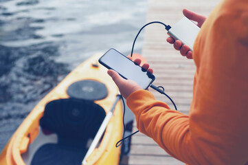 The guy holds a portable charger with a smartphone in his hand. A man against the background of water and a kayak charges the telephone with Power Bank. Concept on the theme of tourism.