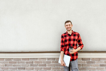 Young happy man freelancer is standing near a bricks wall and holding a coffee and a laptop in his hands