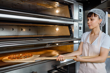 Fototapeta na wymiar Young caucasian woman baker is holding a wood peel with fresh pizza and put it in an oven at a baking manufacture factory.