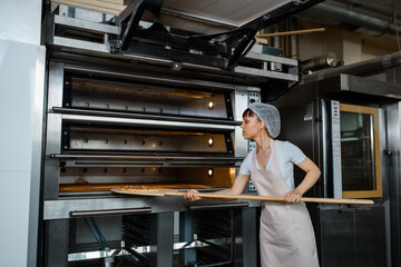 Fototapeta na wymiar Young caucasian woman baker is holding a wood peel with fresh pizza and put it in an oven at a baking manufacture factory.