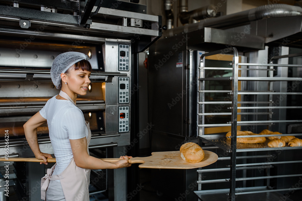 Wall mural young caucasian woman baker is holding a wood peel with fresh bread near an oven at baking manufactu