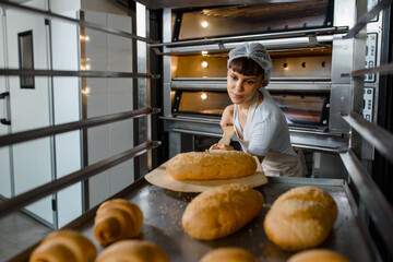 Close up of young caucasian woman baker putting the fresh bread on the shelves/rack at baking manufacture factory.