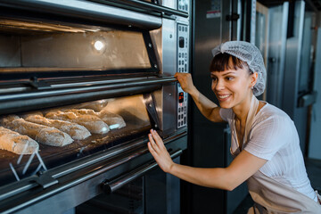 Young caucasian woman baker is looking at the bread baker process in an electric oven at a baking...