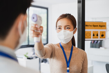 Asian businesswoman wear protective face mask using infrared thermometer check temperature of working people before enter ing office, reopen business with new normal and social distancing concept