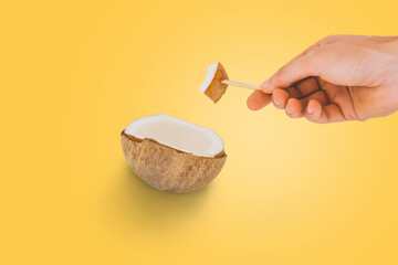 Men's hand holds a piece of pulp of cracked coconut on a wooden stick isolated on a yellow background. A part of fruit. Healthful food. Stylish photograph. Organic food