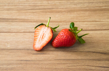 Fresh sweet cut red strawberries with green leaves lies on background of a wooden texture board. One full strawberry lying beside the second half cut strawberry