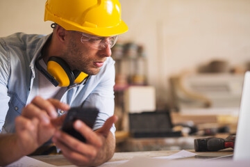 Close up of young carpenter using mobile phone while looking at laptop