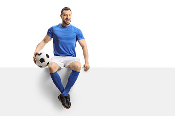 Footballer in a blue jersey with a soccer ball sitting on a blank panel and smiling