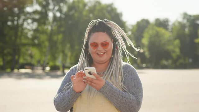 Smiling happy woman with dreads texting message standing at city street during summertime. Stout girl or mobile user looking at mobile screen surfing in internet. Lifestyle and social media concept