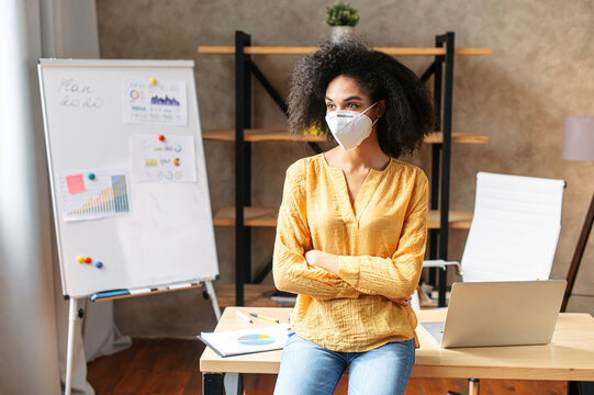 A biracial woman with a protective medical mask on her face standing in the office, a flip chart on the background. Precautionary measures for office employees during pandemic, epidemic