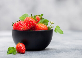 Organic fresh raw strawberries with leaf in black ceramic bowl on light background.