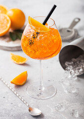Glass of aperol spritz summer cocktail with oranges and strainer with bar spoon and cocktail shaker on light background. Ice cubes and black straw.