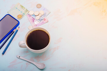 Coffee cups and spoons placed on the table along with a pencil smartphone. With bank notes and coins Laid on the table with the light of the orange sun coming in