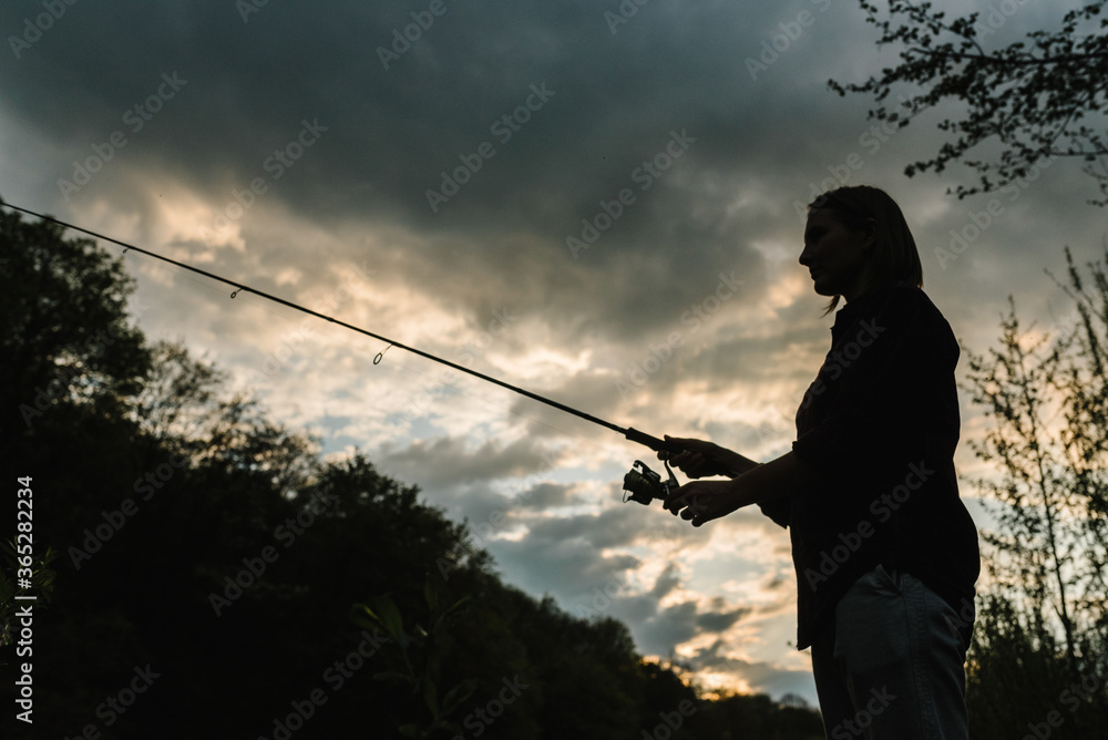 Wall mural Silhouette of a fisherman. Young woman fishing on a lake at sunset. Fishery, fishing day. Rod rings, fishing tackle. Fisherman with rod, spinning reel on the river bank. Fishing for pike, perch, carp.