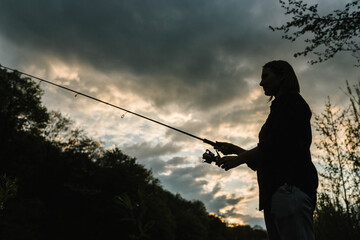 Silhouette of a fisherman. Young woman fishing on a lake at sunset. Fishery, fishing day. Rod rings, fishing tackle. Fisherman with rod, spinning reel on the river bank. Fishing for pike, perch, carp.