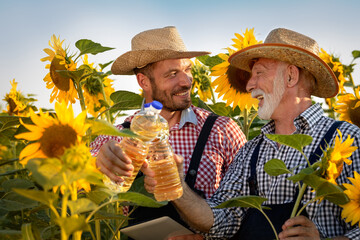 Smiling senior man holding bottle sunflower oil stands in the field with son