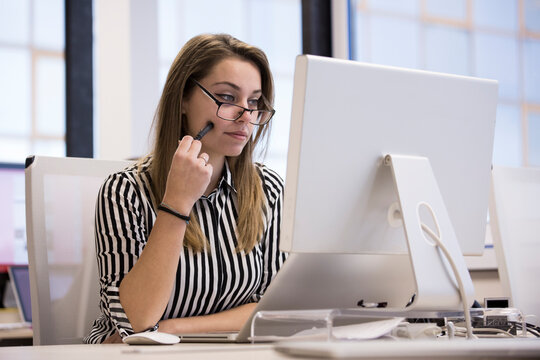 Giovane impiegata  bionda in camicia a righe seduta nella sua postazione lavorativa del suo ufficio lavora di fronte  al computer indossando delle cuffie
