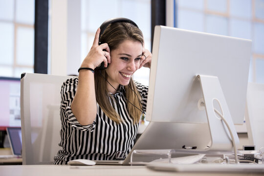 Giovane impiegata  bionda sorridente , in camicia a righe seduta nella sua postazione lavorativa del suo ufficio lavora al computer indossando delle cuffie