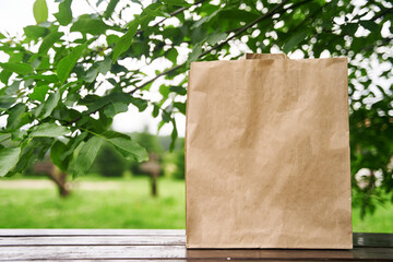 Blank paper bag stands on a wooden table against the background of green leaves. Shopping concept. High-quality photo.