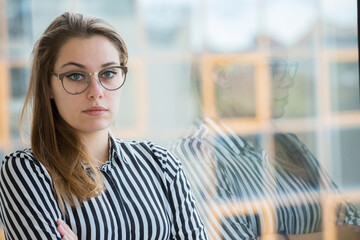 Ragazza bionda di pelle chiara occhiali da vista e una camicia a righe sta in piedi con le braccia conserte vicino alla vetrata del  suo ufficio  da cui si vedono altri edifici