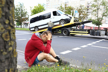 Man sits on side of road holding his head with his hands next to wrecked car after car accident....