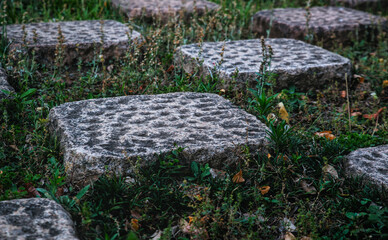 Sidelong perspective of a checkered pattern of dimpled stone tiles and square patches of grass with bits of leaf litter visible