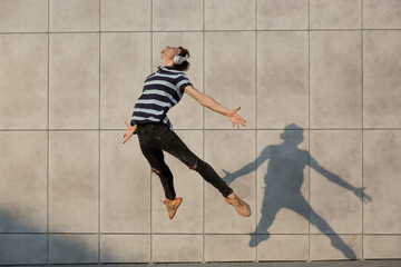Jumping young buinessman in front of city building wall, on the run in jump high. Listening to music, moving to daily routine inspired and sportive. Young ballet dancer in casual clothes and sunshine.
