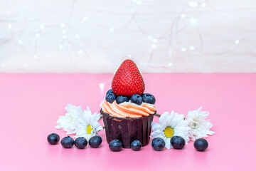 festive little cupcake with cream on a pink background decorated with bright lights, blueberries, strawberries and flowers, close-up
