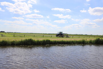 
A farmer is haying with his tractor in his pasture on the waterfront, beautiful panoramic view over Dutch meadows with a beautiful sky in summer.