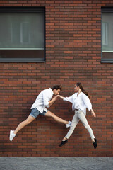 Kissing, greeting. Jumping young couple in front of city building wall, on the run in jump high. Hurrying up, moving to daily routine inspired and sportive. Young ballet dancers in casual clothes.