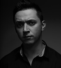 Serious young man looking suspiciously in black shirt on studio dark shadow background. Closeup portrait.