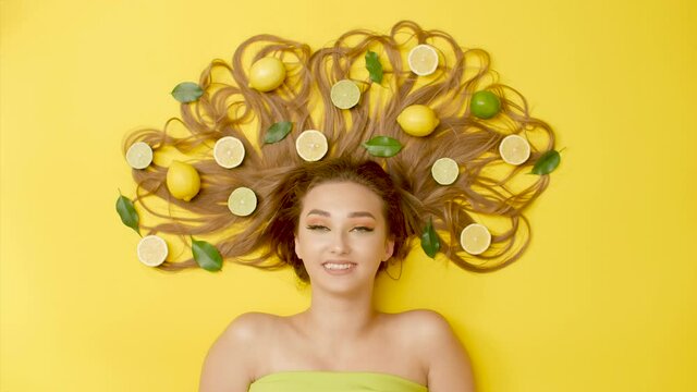funny girl lying on yellow background with citrus fruits on long hair dancing with eyebrows , young woman flirting and have fun, concept female mood,head with lemons