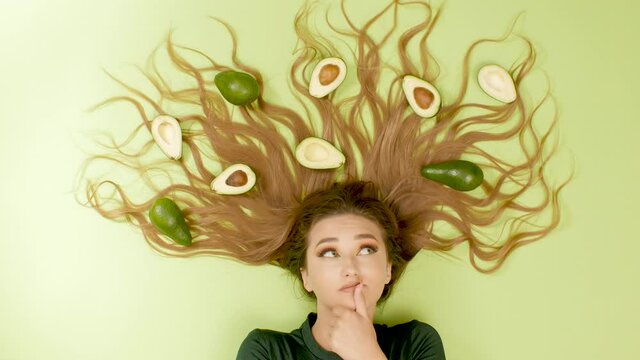 beautiful girl lying on green colored background with avocado fruits on long hair, young woman thinking with hand under chin, concept choice balance