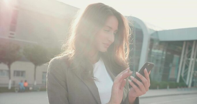 Pretty woman with Long Brown hair and Nice Hairstyle Looking at the while typing on her Smartphone. Classically dressed. Standing by a big Business building. Sun rays going on the camera.