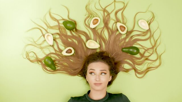 beautiful girl lying on green colored background with avocado fruits on long hair, young woman dancing with eyebrows, concept healthy lifestyle, nutrition, energy, beauty