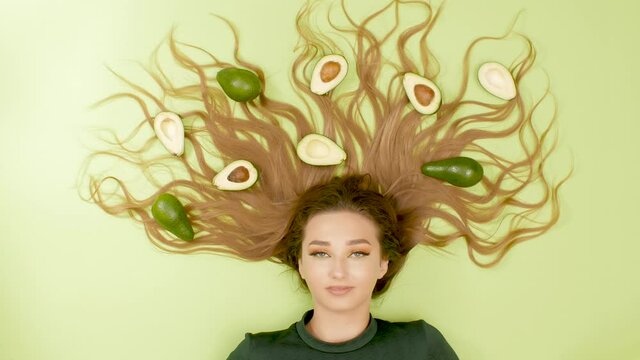 beautiful girl lies on bright colored background with avocado fruits on long hair, young woman dances with eyebrows, concept creative beauty idea, female positive mood