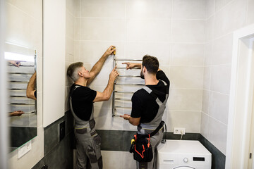 Two plumbers, workers in uniform working, installing towel rail, electric towel dryer on the...
