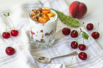 Breakfast parfait with yogurt, granola, peach and sour cherries in a glass. Vintage silver spoon and fruit in the background