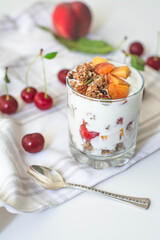 Breakfast parfait with yogurt, granola, peach and sour cherries in a glass. Vintage silver spoon and fruit in the background