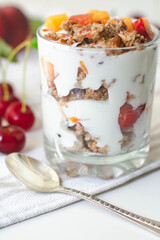 Breakfast parfait with yogurt, granola, peach and sour cherries in a glass. Vintage silver spoon and fruit in the background