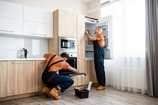 Safe and affordable repairs. Two handymen, workers in uniform fixing, installing furniture and equipment in the kitchen, using screwdriver indoors. Furniture repair and assembly concept
