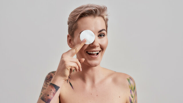 Portrait of beautiful tattooed woman with pierced nose and short hair smiling while holding cotton pad for removing makeup, cleaning skin isolated on grey background