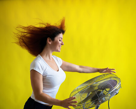 A Beautiful Red-haired Woman Is Cooled Off Standing Over A Large Electric Fan On A Yellow Background. Girl With Hair Developing In The Wind. Device For Cooling The Air.