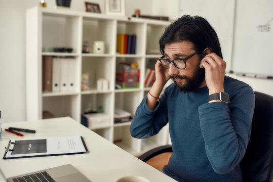 Close up portrait of young bearded male teacher adjusting wireless headphones while giving online class through webcam at home