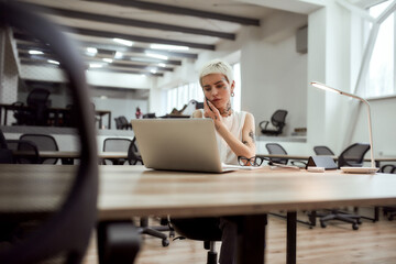 Important talk. Young and beautiful tattooed business woman talking on mobile phone and using laptop while sitting at her working place in the modern office