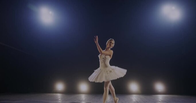 Camera zooming in young pretty hard-working graceful ballerina performing classical ballet pas on stage of theater or studio in blue lamps light. Choreography concept.